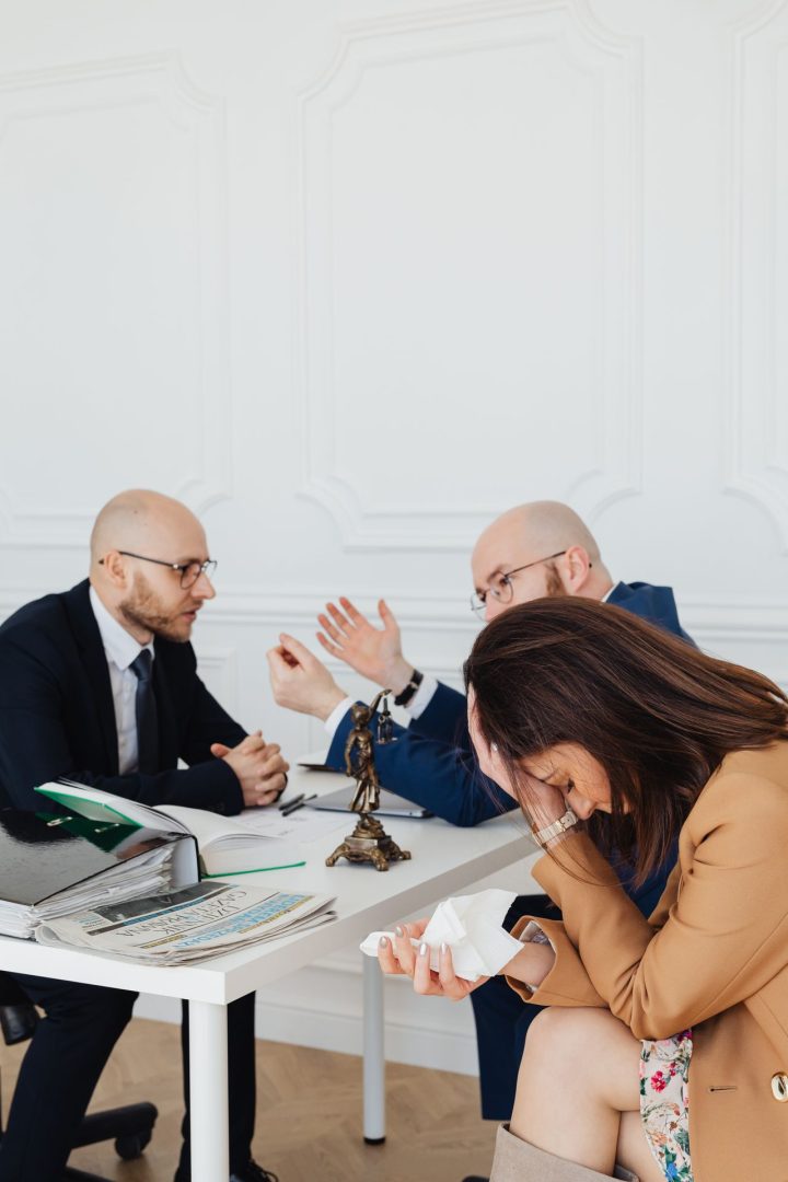 A woman sits at a desk crying into a tissue, while two men in suits have a serious discussion in the background, with legal documents and a small justice statue on the table.