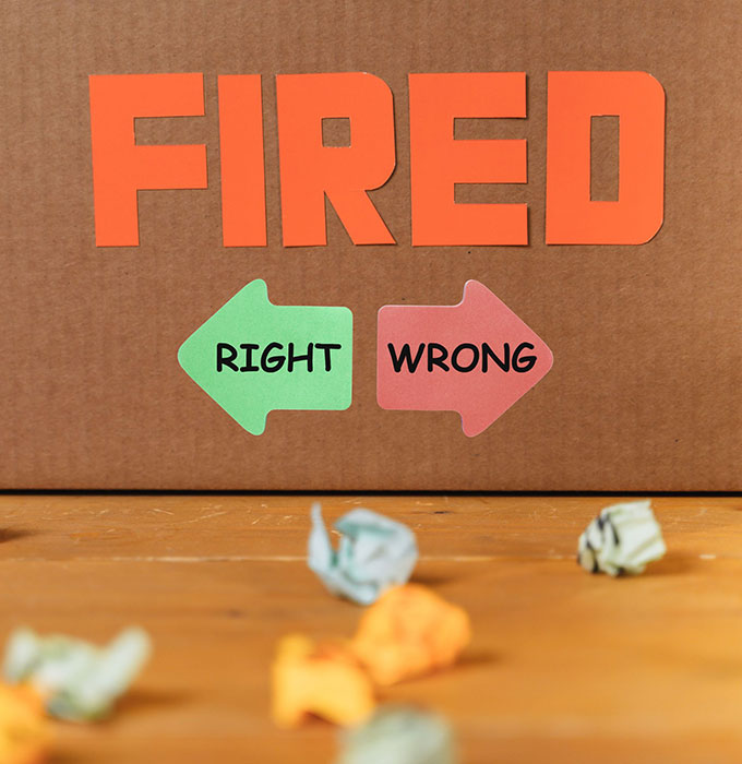 A cardboard sign with large orange letters spelling "FIRED" above two arrows: a green one labeled "RIGHT" and a red one labeled "WRONG." Crumpled paper balls are scattered on a wooden surface below.