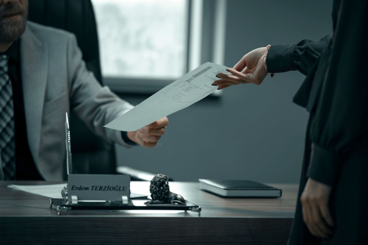 A person hands a document to another person seated at a desk with a nameplate, notebook, and pen holder visible. The scene appears to be in an office setting.