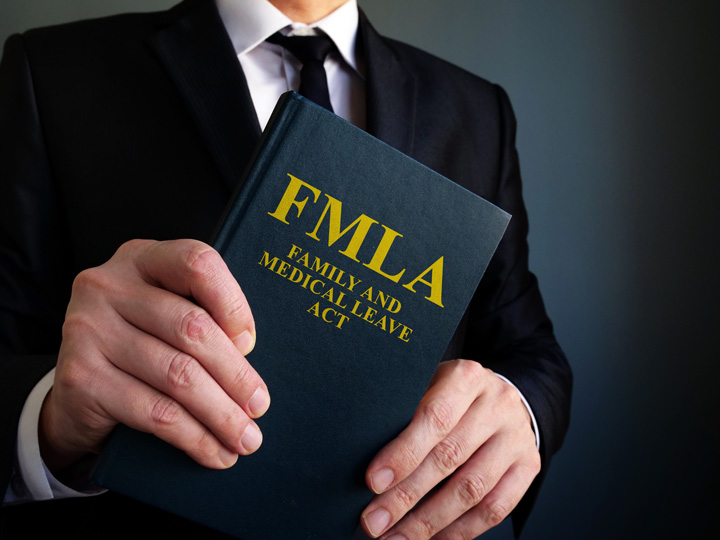 A person in a suit holds a book titled “FMLA Family and Medical Leave Act” against a plain dark background.