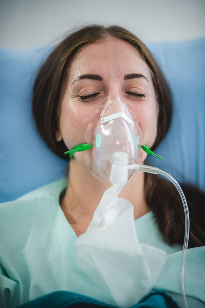 A woman with closed eyes lies in a hospital bed, wearing a nasal oxygen mask and a light green gown, appearing to rest or recover.