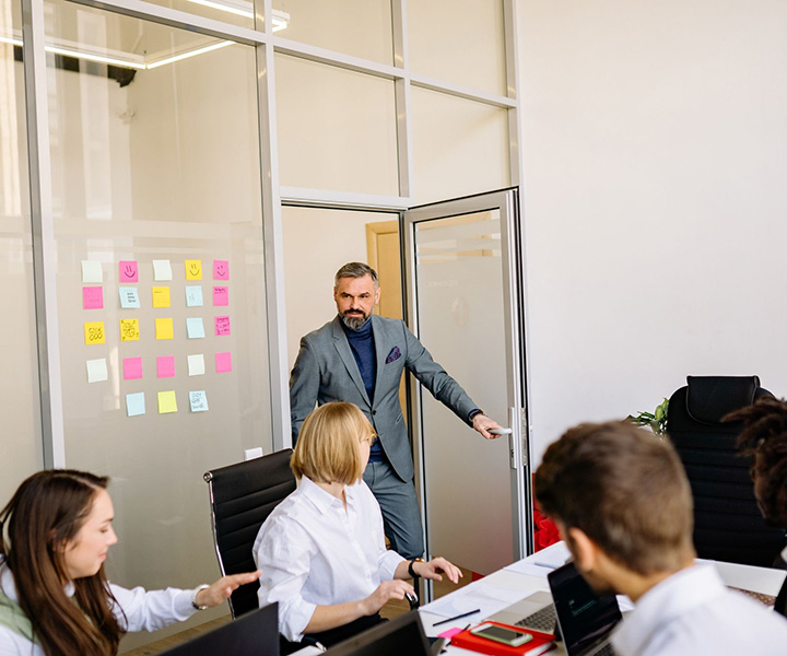 A man in a suit enters a conference room where four people are seated at a table, discussing and working on laptops. Colorful sticky notes are visible on glass walls in the background.