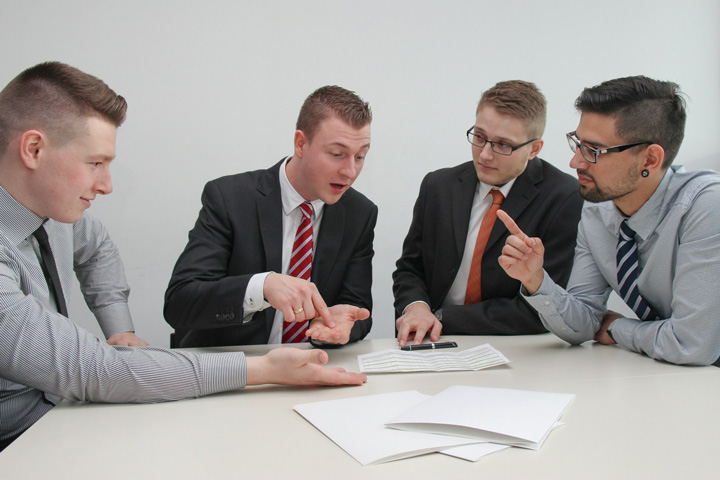 Four men in business attire sit at a table discussing documents. Two men gesture with their hands while the other two listen. Papers are spread out in front of them. The background is plain and white.